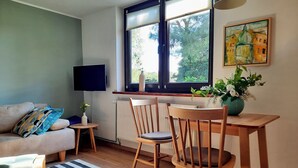 Ein Wohnzimmer mit einer Couch, einem Esstisch mit Stühlen, einem Fernseher, einem Fenster mit Jalousien und einem Bild an der Wand.