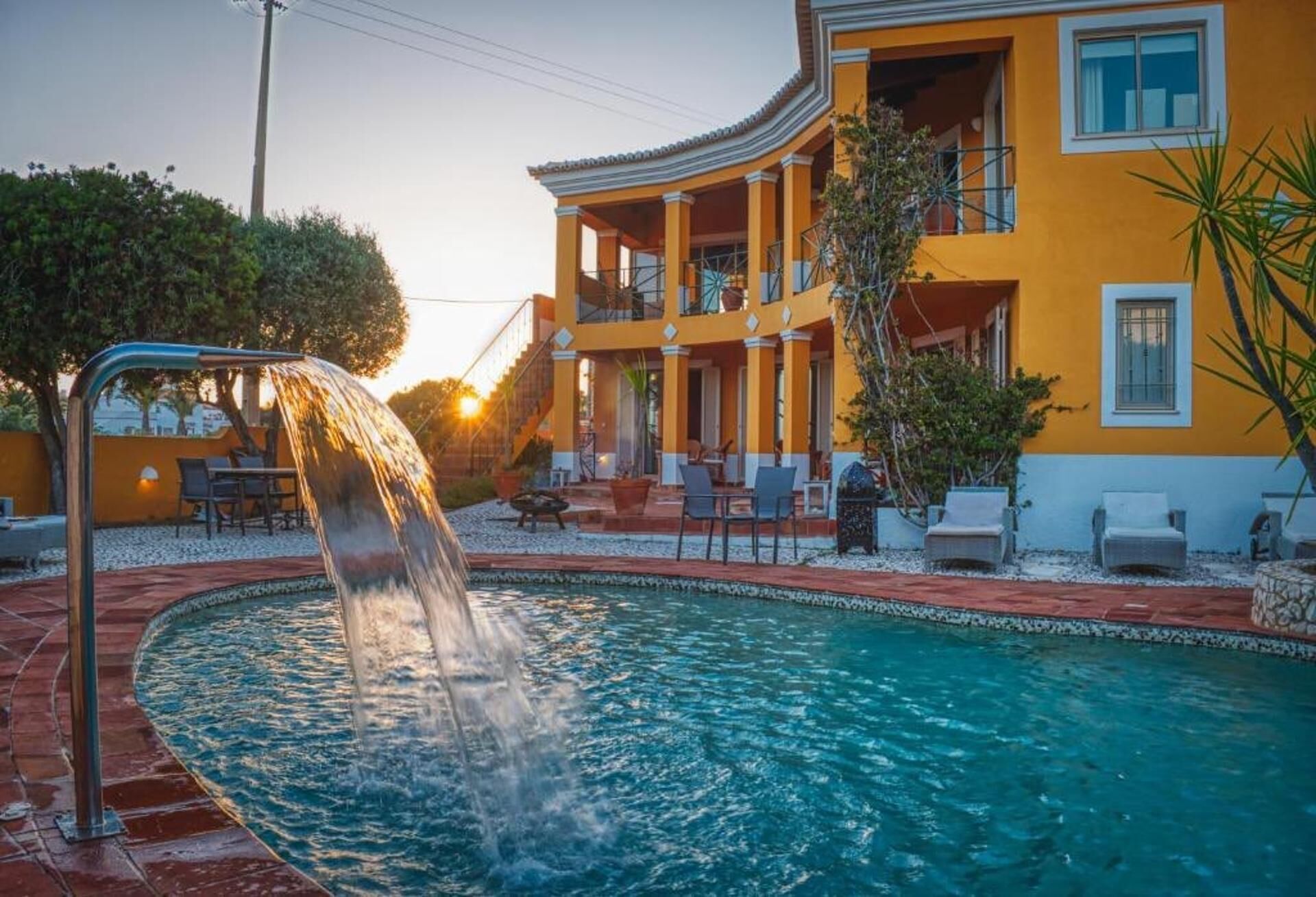 A pool with a water feature, a yellow building with balconies, and outdoor seating.