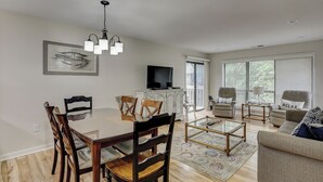 Un comedor con mesa de vidrio y sillas de madera, una sala de estar con sofá y sillones, un televisor montado en la pared y un ventanal con persianas.