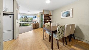 Ein Essbereich mit Holztisch und -stühlen, eine Küche mit einem weißen Kühlschrank und Blick durch eine Glastür auf einen Außenbalkon.