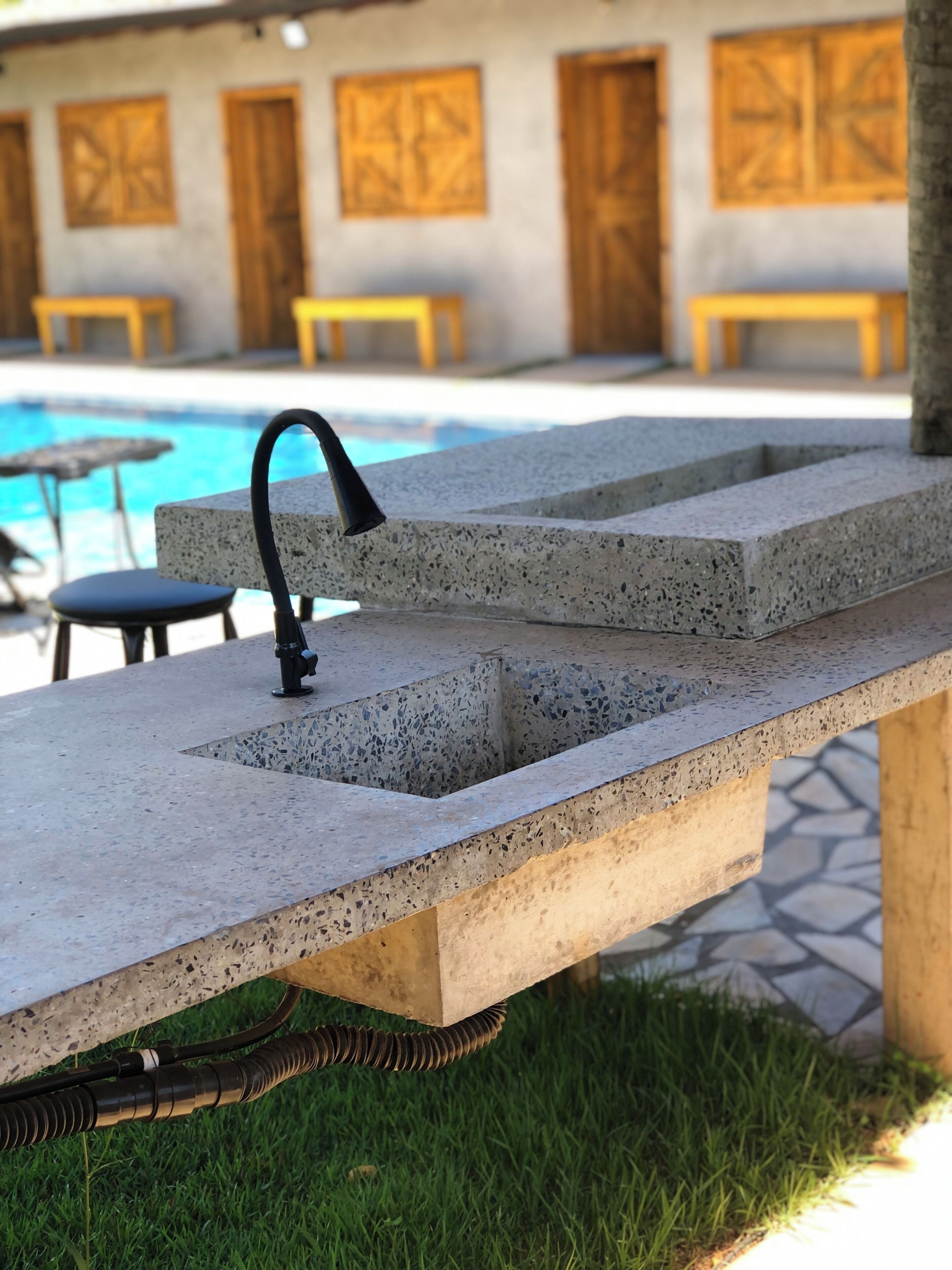 Una barra exterior de hormigón con un grifo negro, una piscina al fondo y suelo de baldosas de piedra.