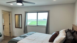 A bedroom with a bed, a ceiling fan, a window with a view of a field, and a door.