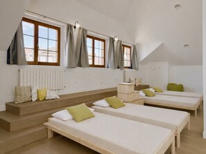 Hotellihuone, jossa on kolme erillistä sänkyä, puinen penkki, patteri ja ikkuna, jossa on verhot.