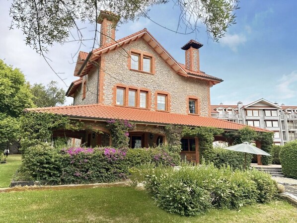 A stone building with a red-tiled roof, multiple windows, and a covered porch with a garden in front. - Villa Las tres Mares 6826 (Noja)
