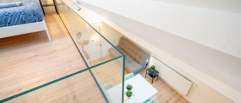 A bedroom with a bed, a glass-walled balcony, a sofa, a table with chairs, and a radiator.
