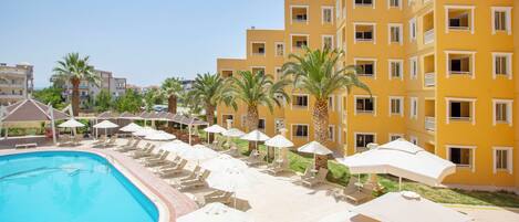 A hotel with a swimming pool, sun loungers, umbrellas, and palm trees.