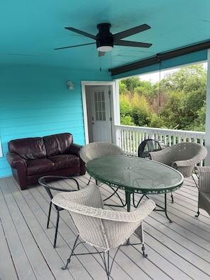 Eine überdachte Veranda mit Deckenventilator, einem grünen Metalltisch, Korbstühlen, einem braunen Ledersofa und einer weißen Tür.