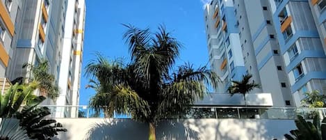 A modern high-rise building with multiple balconies, a palm tree in the foreground, and a scooter parked on the street.