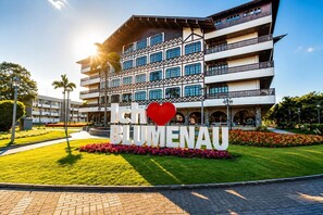 A multi-story hotel with a prominent sign, a landscaped garden, and a clear sky. - Bem-estar e Segurança, em localizaçao privilegiada. (Blumenau)