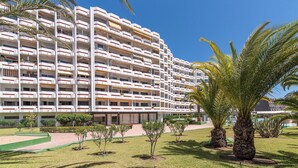 Een hotel met meerdere verdiepingen, balkons, een aangelegde tuin met palmbomen en een helderblauwe lucht.