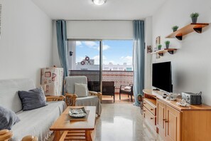 Ein Wohnzimmer mit Sofa, Sesseln, Fernseher, Holzsäcken und einem Balkon mit Stadtblick.