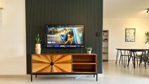 A modern living room with a flat-screen TV mounted on a wooden cabinet, a dining table, and a potted plant. - Beautiful 5-BR House with AC in Zaragoza. Community Pool & Close to the Beach! (Zaragoza)