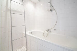 A white tiled bathroom with a bathtub, a shower, and a metal towel rack. - 3-bedroom Apartment in National Residence (Antwerp)