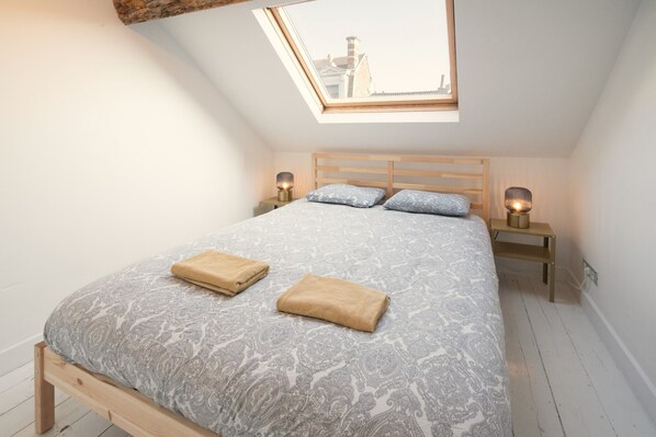 A bedroom with a skylight, a bed with patterned bedding, two bedside tables with lamps, and a wooden headboard. - 3-bedroom Apartment in National Residence (Antwerp)