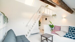 Uma sala de estar moderna com um sofá cinzento, uma pequena mesa de madeira, uma escadaria branca e um teto com vigas aparentes.