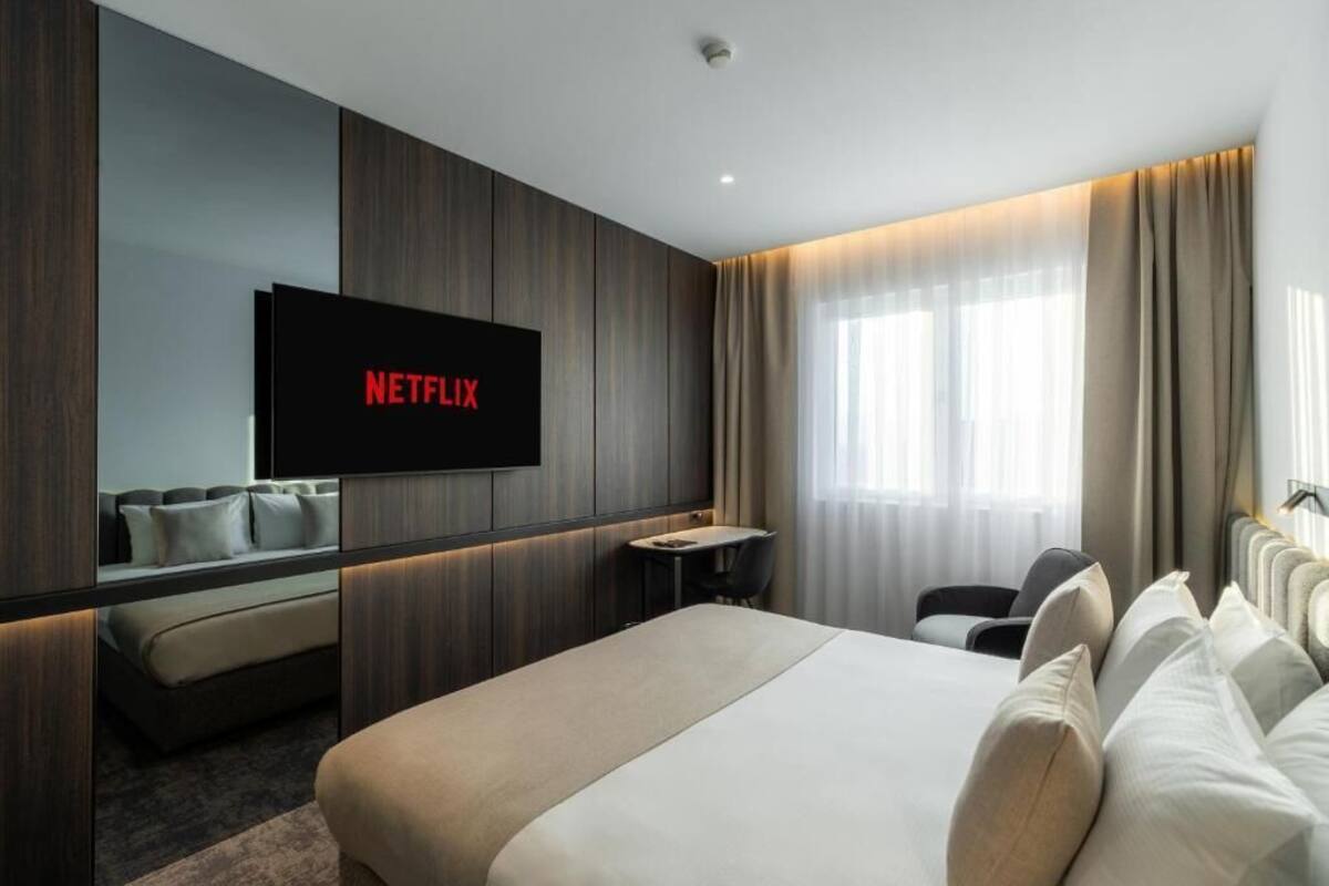 Ein modernes Hotelzimmer mit einem großen Bett, einem Schreibtisch und einem an der Wand befestigten Fernseher, der 'NETFLIX' anzeigt.