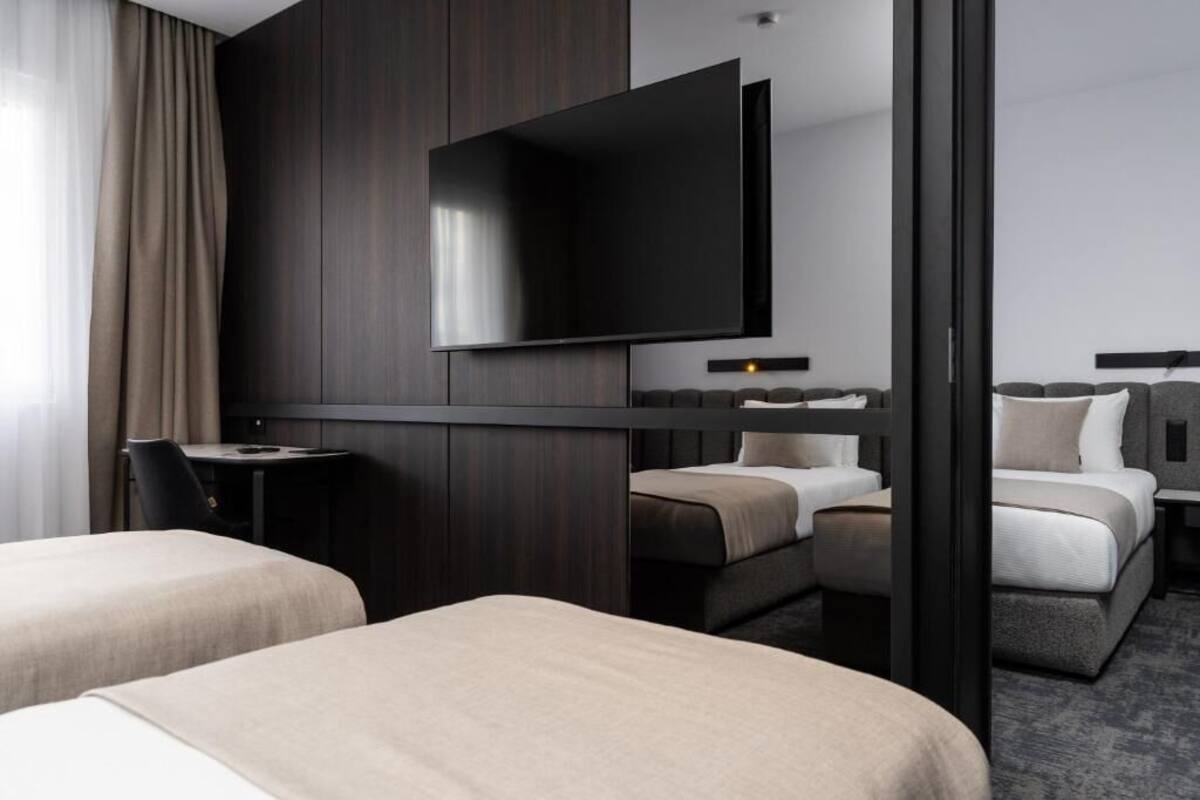 Ein Hotelzimmer mit Flachbildfernseher an der Wand, zwei Betten, einem Schreibtisch und einem Sessel.