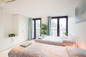 Quarto moderno com duas camas, uma janela grande com cortinas, uma planta em vaso e vista para prédios.