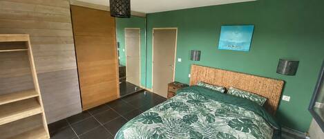 A bedroom with a bed, a wooden wardrobe, a nightstand, a wall-mounted lamp, and a painting.