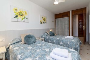 Ein Hotelzimmer mit zwei Betten, blumigem Bettbezug, Deckenventilator, Bildern an der Wand und einer Holztür.