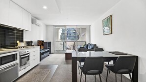 Una cocina moderna con armarios blancos, un comedor con mesa de vidrio y vistas a un edificio a través de amplios ventanales.