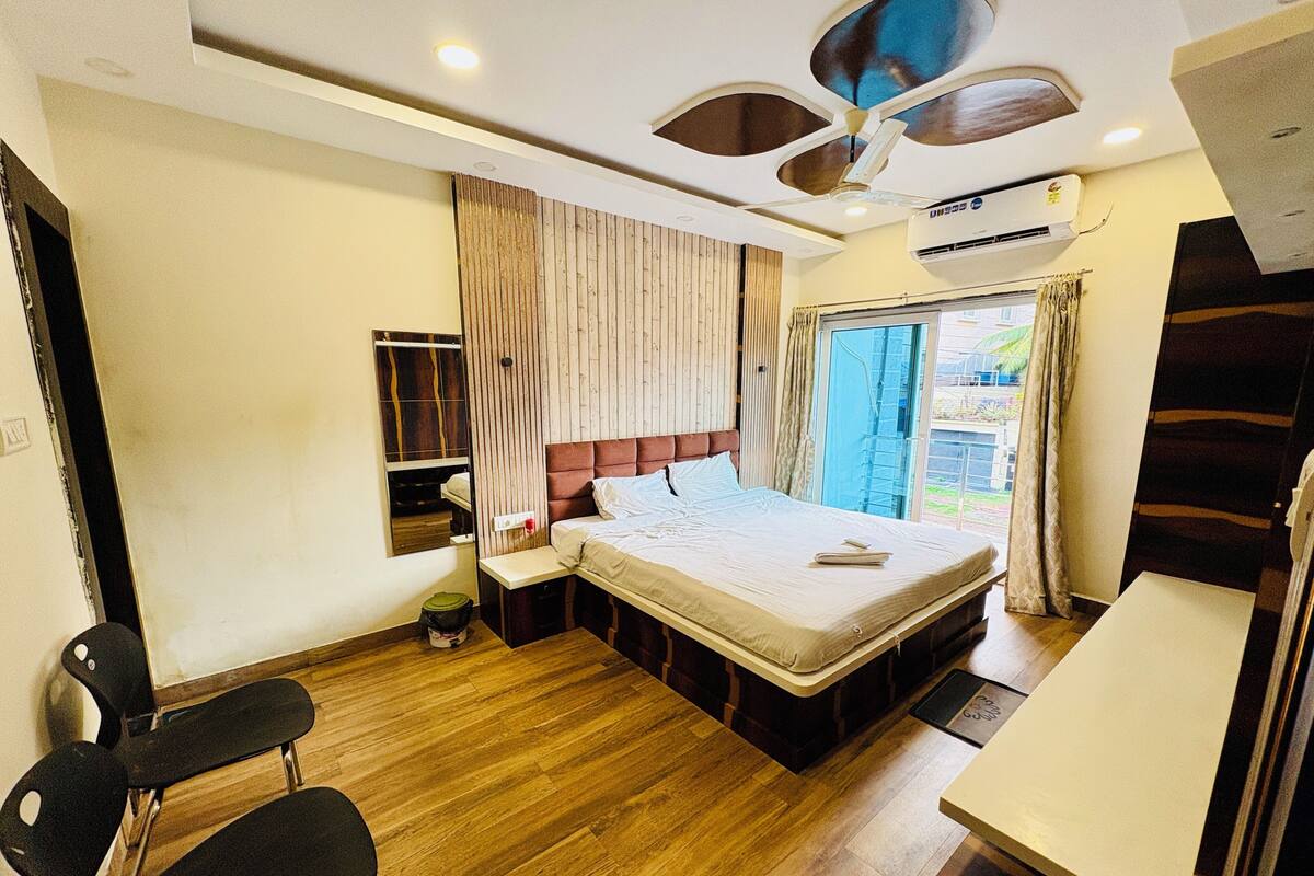 A bedroom with a bed, a chair, a bedside table, a mirror, a ceiling fan, and an air conditioning unit.