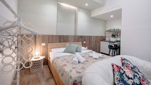 A modern hotel room with a wooden headboard, a bed with floral bedding, a small kitchenette, and a hanging light fixture.