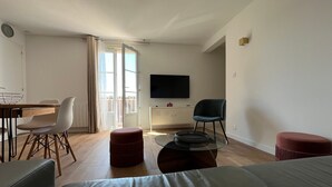 Uma sala de estar moderna com um sofá, uma mesa de jantar com cadeiras, uma TV e uma varanda com cortinas.