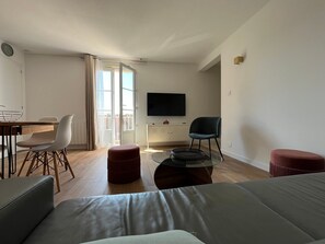 Ein modernes Wohnzimmer mit Sofa, Esstisch mit Stühlen, Fernseher und einem Balkon mit Vorhängen.