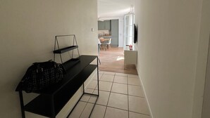 Um corredor com um banco, uma área de refeições com mesa e cadeiras, e uma cozinha ao fundo.