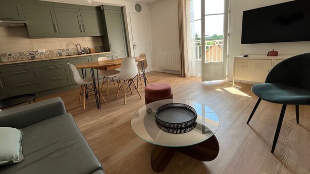 Uma sala de estar moderna com sofá, mesa de centro de vidro, área de jantar com cadeiras, cozinha com armários, televisão e vista para varanda.