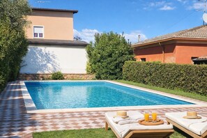 Ein Schwimmbecken mit gefliesten Rändern, umgeben von einer Schachbrett-Terrasse, neben einem Gebäude mit einem gepflasterten Dach.