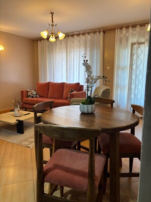 A dining area with a round wooden table, red upholstered chairs, a vase with flowers, and a sofa in the background. - Calm family friendly 2 bedroom apartment in the suburbs of Entebbe (Entebbe)