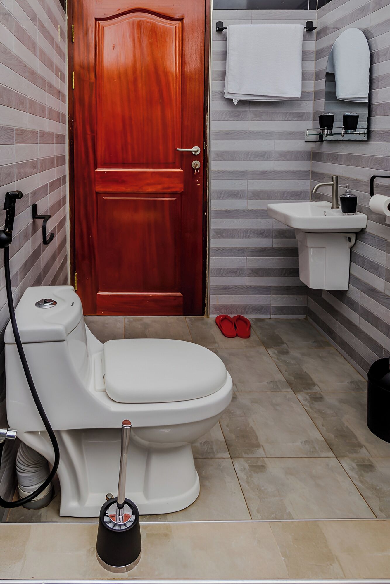 Een badkamer met toilet, wastafel, houten deur en een paar rode slippers op de vloer.