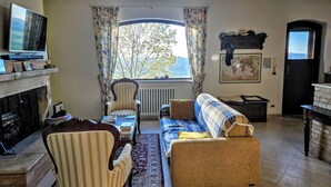 Ein Wohnzimmer mit Kamin, einer Couch, einem Sessel, einem Fernseher und Blick ins Freie.