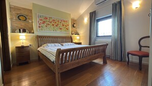 Ein Schlafzimmer mit einem hölzernen Bett, Nachttischen mit Lampen, einem Fenster mit Vorhängen und einem an der Wand befestigten Regal mit dekorativen Gegenständen.