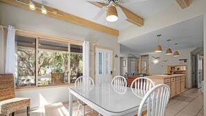 Un coin repas avec une table en verre et des chaises blanches, une cuisine avec des meubles en bois et une vue sur les arbres à travers de grandes fenêtres.