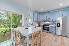 A modern kitchen with light blue cabinets, stainless steel appliances, and a dining area with a white table and wooden chairs. - The Tree House •  Dog Friendly Beach Escape (Oak Island)