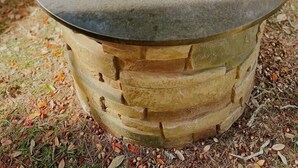Uma lareira de pedra com borda de metal, rodeada por um espaço gramado com um banco e uma cadeira ao fundo.