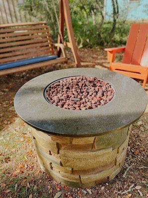 A stone fire pit with a metal rim, surrounded by a grassy area with a bench and a chair in the background. - The Tree House •  Dog Friendly Beach Escape (Oak Island)