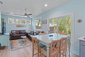A spacious living room with a large dining table, a sofa, and a ceiling fan. - The Tree House •  Dog Friendly Beach Escape (Oak Island)