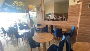 L’intérieur d’un restaurant présente des tables rondes et des chaises bleues, un comptoir avec un menu, et un sol carrelé.
