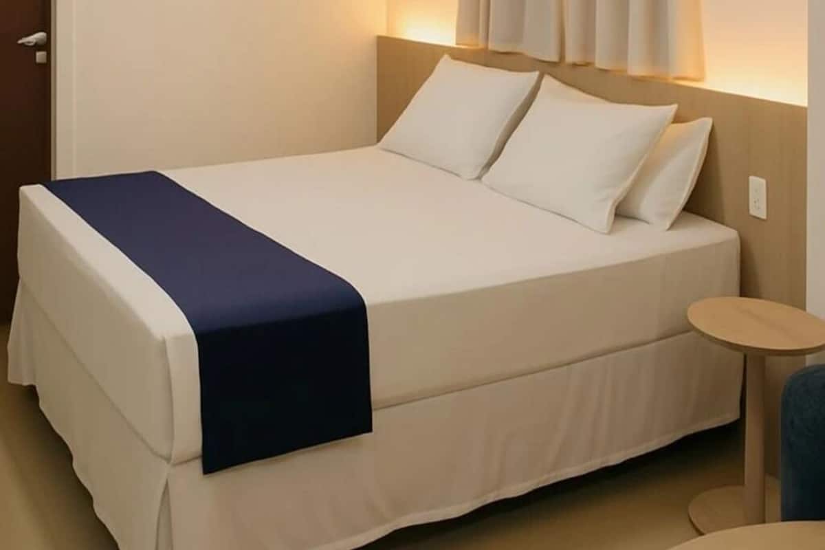 A neatly made bed with a blue bedspread, a small round table, and a wall-mounted light fixture.