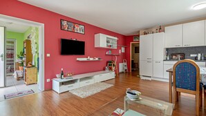 Un soggiorno con parete rossa, pavimento in legno, TV a parete, mobile TV bianco e angolo cottura con pensili e forno a microonde.