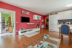 A living room with a red wall, wooden flooring, a TV mounted on the wall, a white entertainment unit, and a kitchen area with cabinets and a microwave.