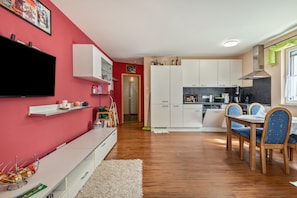 Une cuisine moderne avec des meubles blancs, un coin repas avec des chaises en bois et un téléviseur à écran plat fixé à un mur rouge.