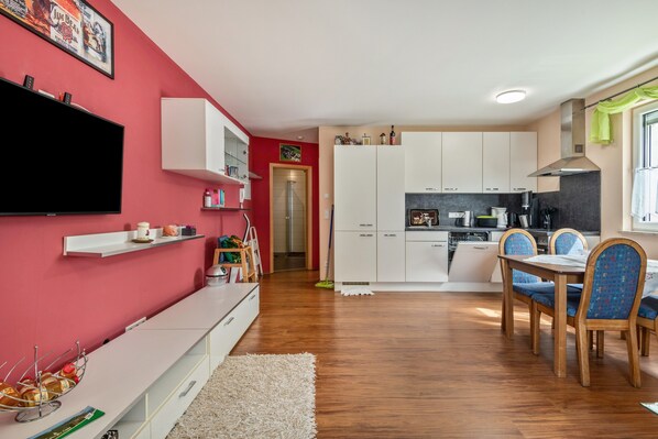 Eine moderne Küche mit weißen Schränken, ein Essbereich mit Holzstühlen und ein Flachbildfernseher an einer roten Wand.