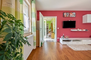 Une pièce avec un plancher en bois, une console blanche et un téléviseur fixé au mur, ainsi qu’une plante dans un coin.