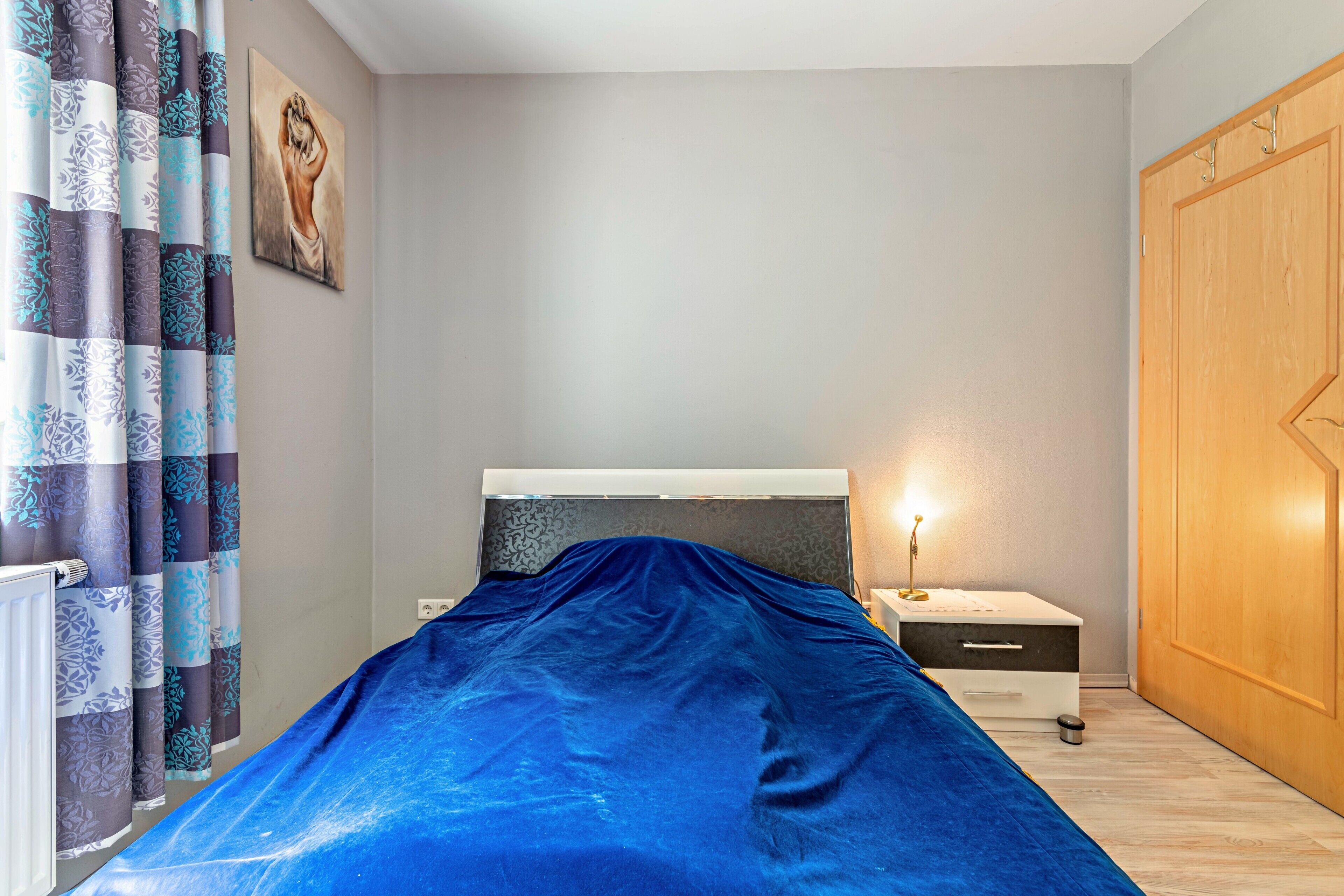 A bedroom with a bed covered in blue, a nightstand with a lamp, a wooden wardrobe, and patterned curtains.