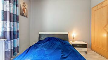 A bedroom with a bed covered in blue, a nightstand with a lamp, a wooden wardrobe, and patterned curtains.